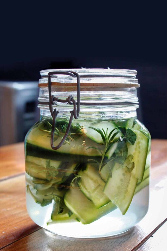 A small glass jar filled with sandwich pickles, rosemary, and thyme.