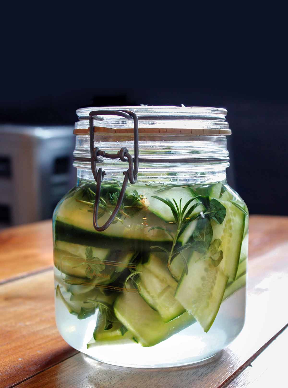 A small glass jar filled with sandwich pickles, rosemary, and thyme.