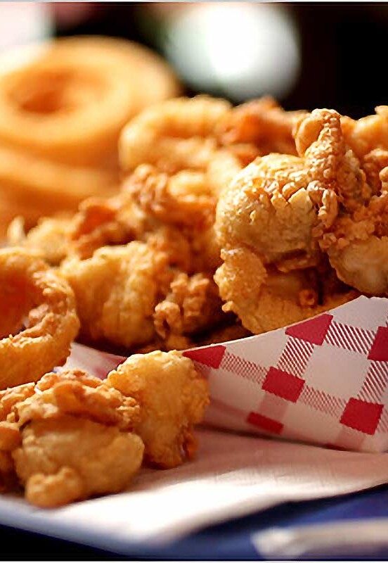 A red and white paper container of whole-belly fried clams, behind is onion rings and condiments