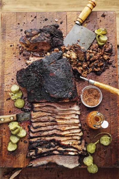 A partially sliced barbecue beef brisket on a cutting board with spice rub, hot sauce, and pickle slices scattered around it.