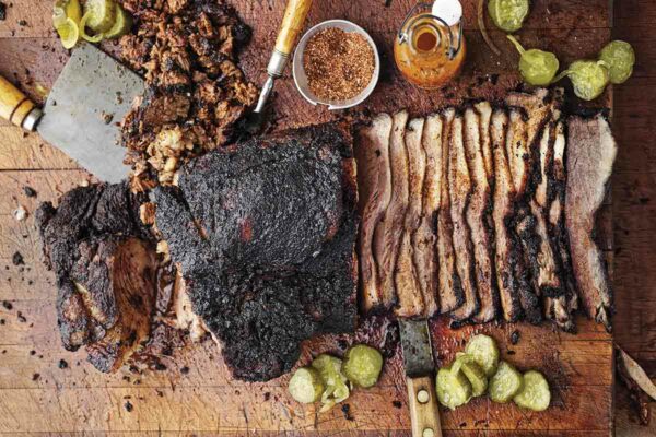A partially sliced barbecue beef brisket on a cutting board with spice rub, hot sauce, and pickle slices scattered around it.