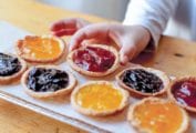 A tray of mini jam tarts--orange, strawberry, blueberry--one being picked up by a child's hand.