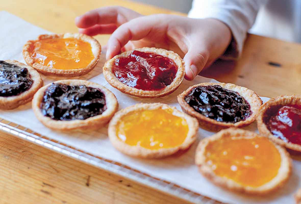 A tray of mini jam tarts--orange, strawberry, blueberry--one being picked up by a child's hand