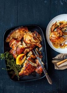 A black platter with golden brown herb butter chicken, roasted onion, a lemon wedge, and sprigs of thyme and oregano by Dorie Greenspan