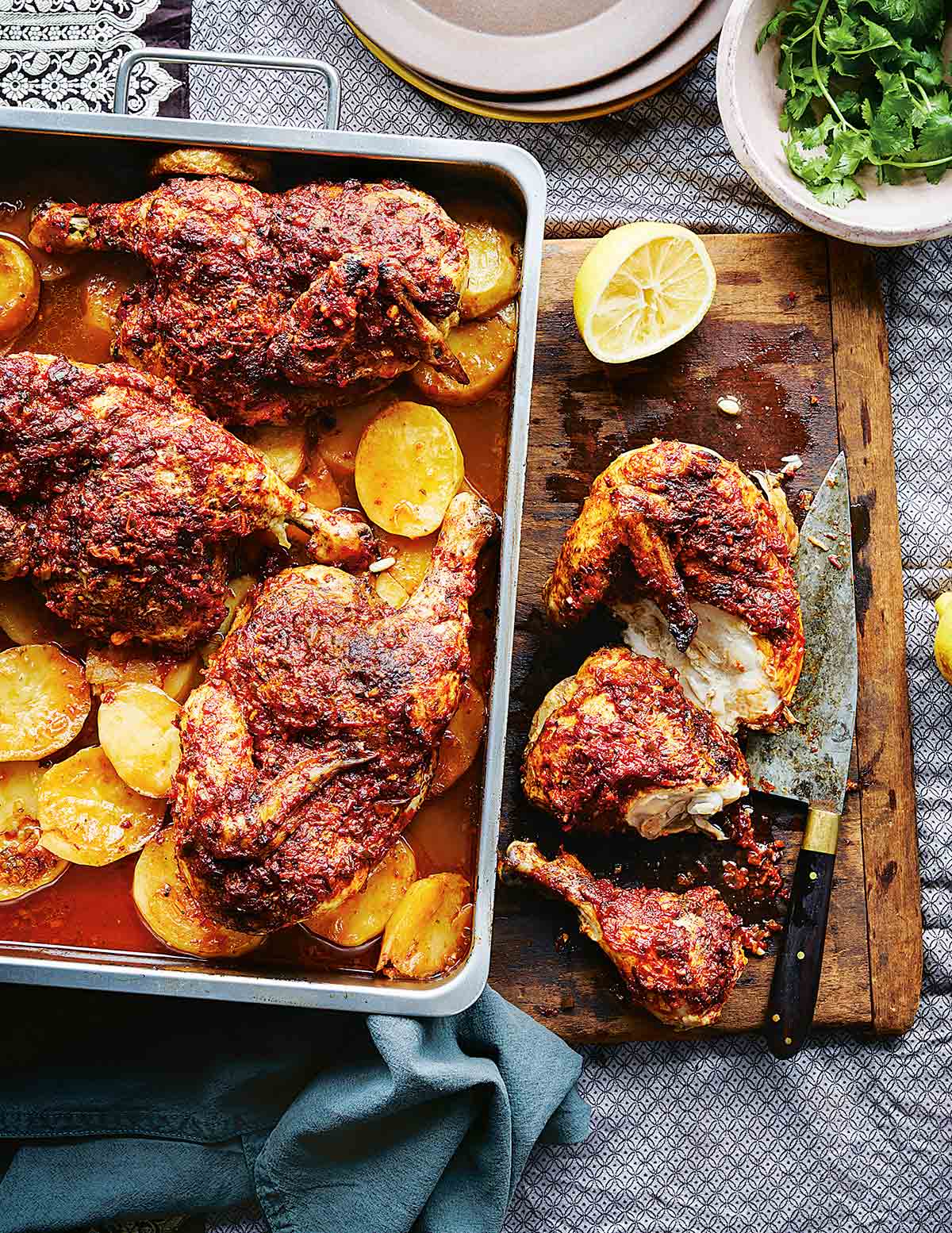 A pan of harissa roasted chicken sitting on top of sliced roasted potatoes, nearby a cutting board and knife.