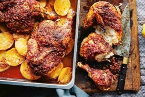 A pan of harissa roasted chicken sitting on top of sliced roasted potatoes, nearby a cutting board and knife