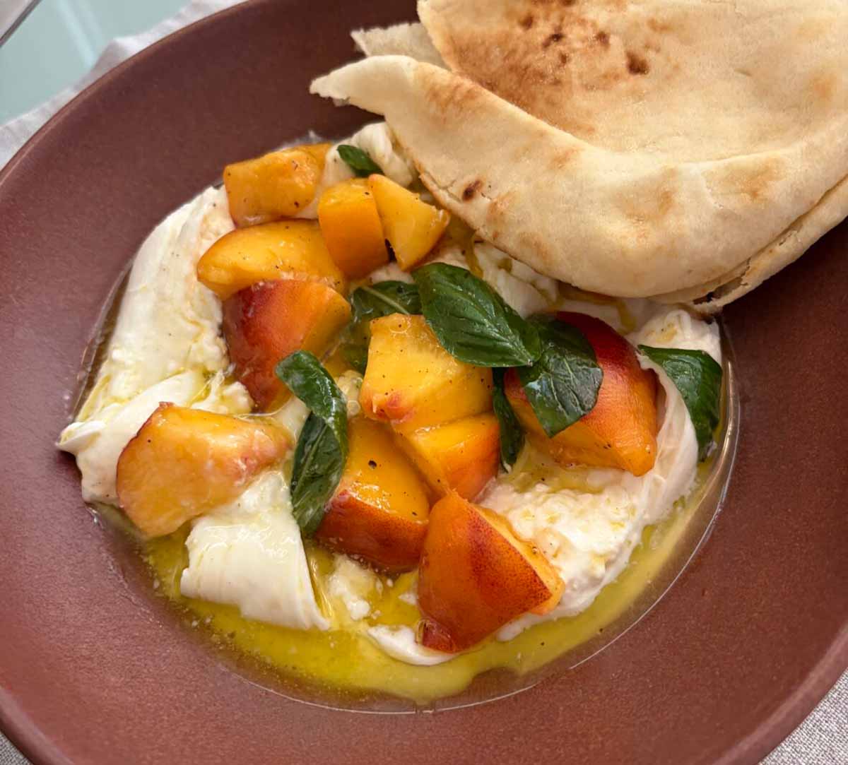An ochre bowl filled with Barata, fresh peaches, fresh basil, and a drizzle of olive oil.