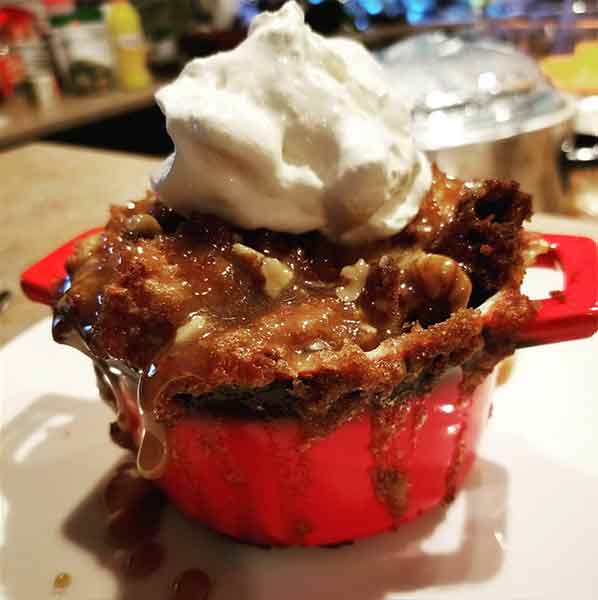 Small red casserole pot frilled with pecan pie bread pudding with caramel dripping off the sides and topped with shipped cream
