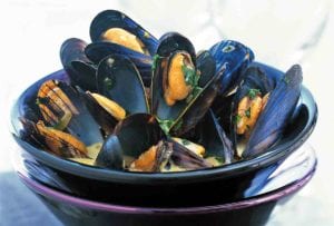 A bowl of pressure cooker mussels with saffron sprinkled with chopped parsley and in a yellow broth set in another bowl on top of a napkin