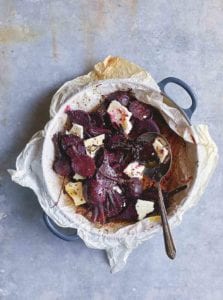 A pan lined with parchment paper containing sliced roasted beets, chunks of feta cheese, a dressing, and a spoon.
