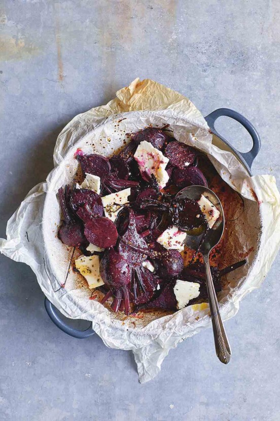 A pan lined with parchment paper containing sliced roasted beets, chunks of feta cheese, a dressing, and a spoon.