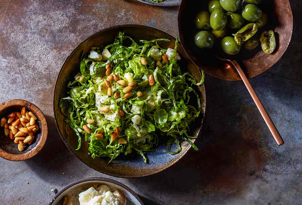 Two plates of shredded, shaved Brussel sprouts salad, topped with toasted pine nuts, green olives, bread, and more pine nuts nearby