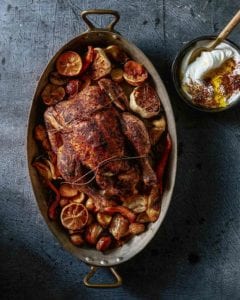 An oval pan filled with a whole roast chicken with sumac, surrounded by roasted carrots, garlic, lemon wedges; a bowl of yogurt, sumac, and olive oil nearby