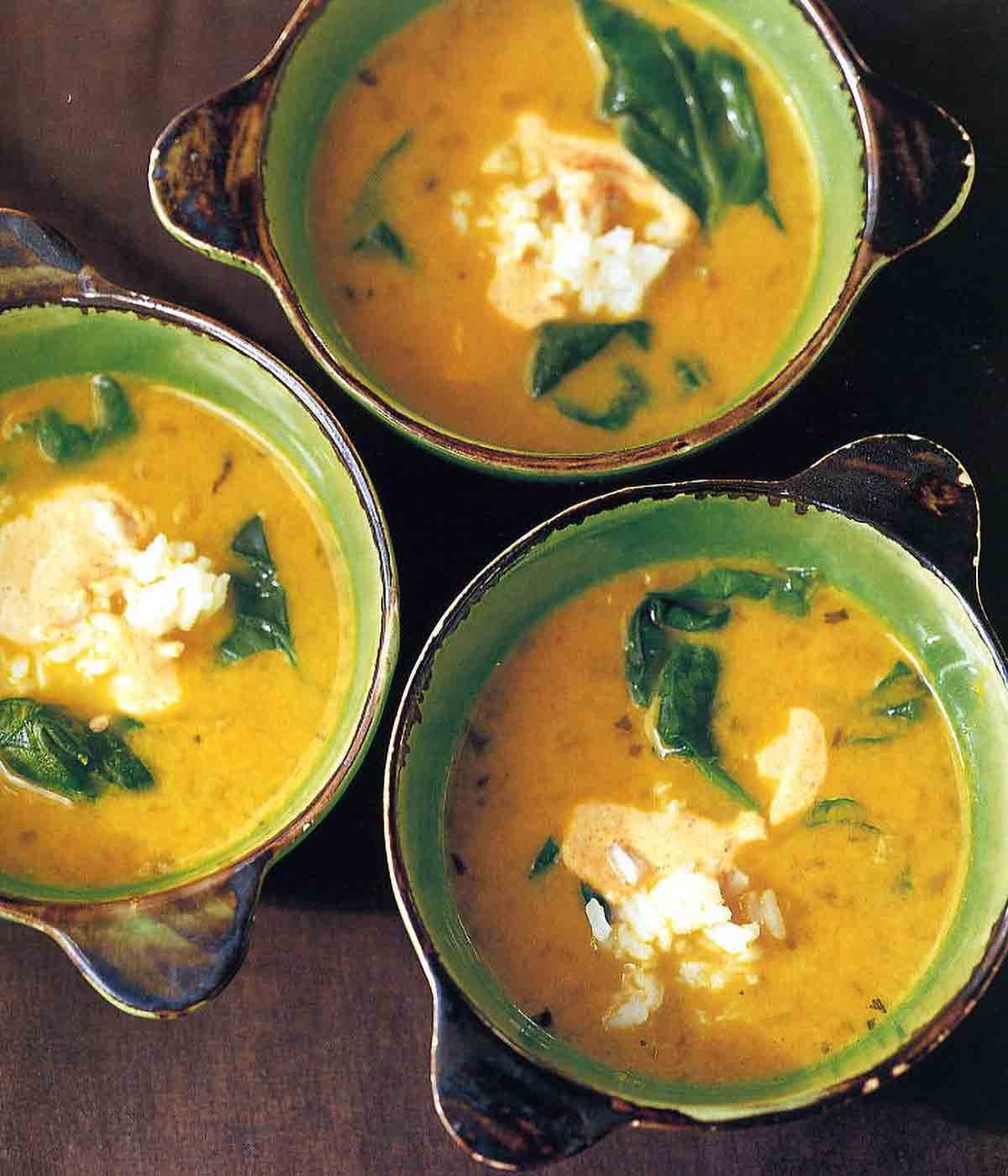 Three bowls of yellow pea and coconut milk soup with pieces of spinach and a dollop of yogurt in each bowl.