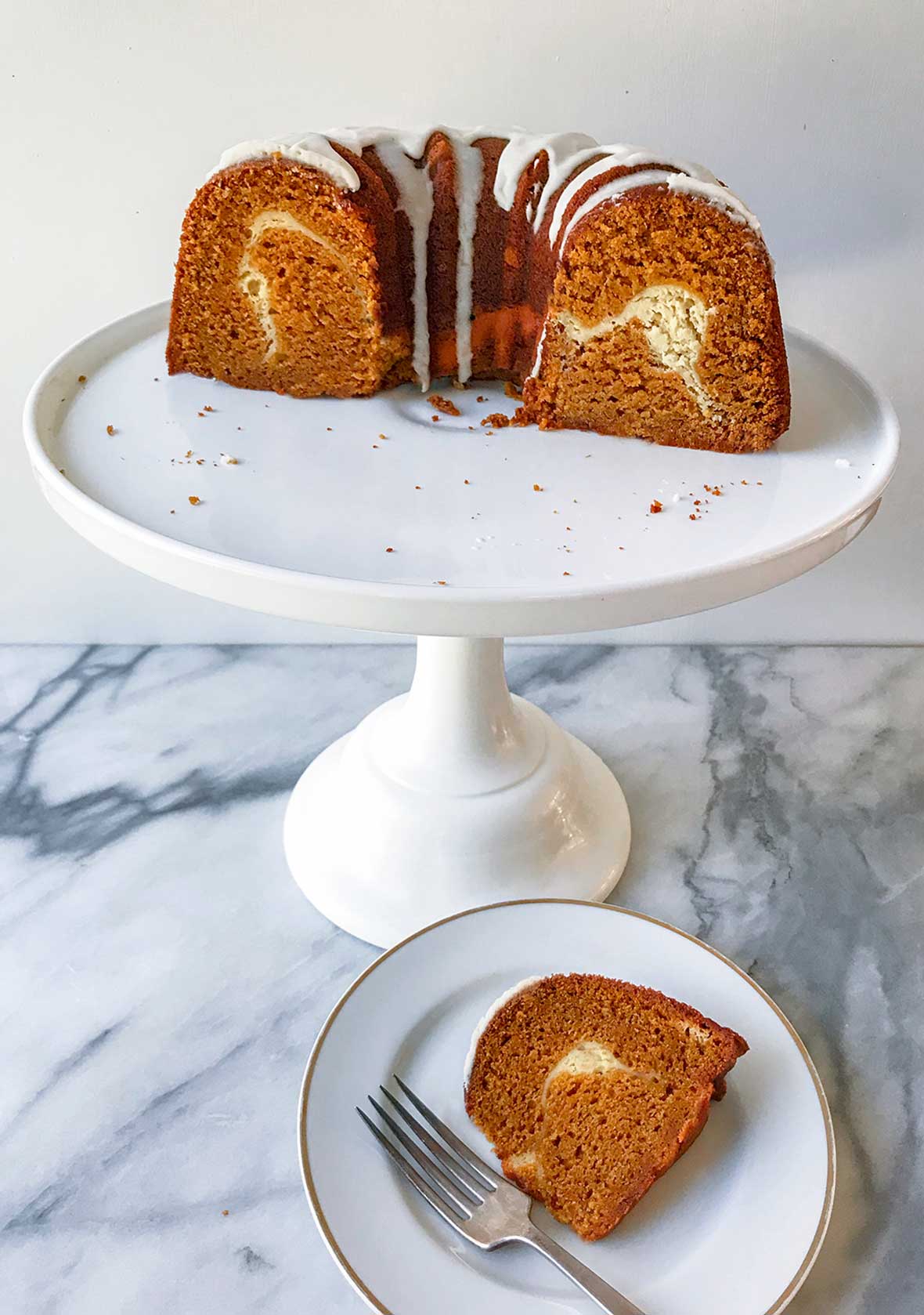Half of a glazed pumpkin pound cake with a swirl of cream cheese filling inside on a cake stand, a piece on a plate nearby