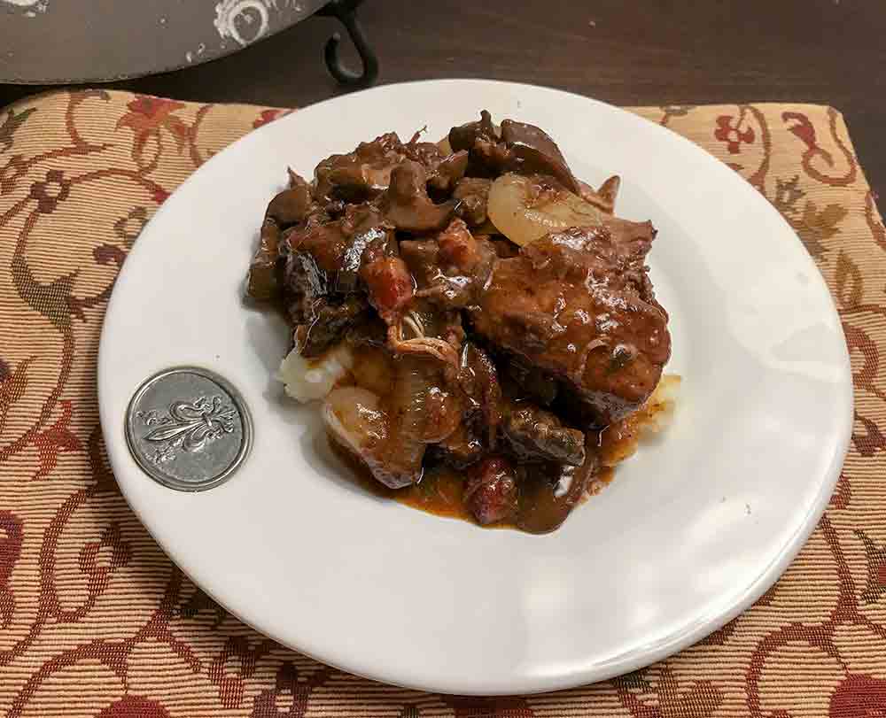 White plate with a silver crest filled with Julia Child's coq au vin--pieces of chicken in red wine, onions, mushrooms, bacon, over mashed potatoes