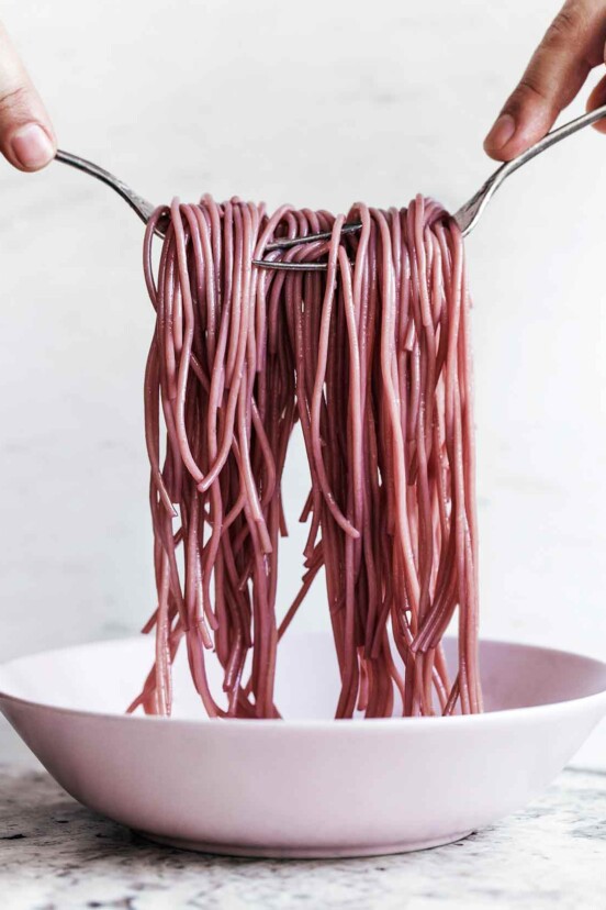 A person using two forks to lift strands of red wine spaghetti out of a white bowl.