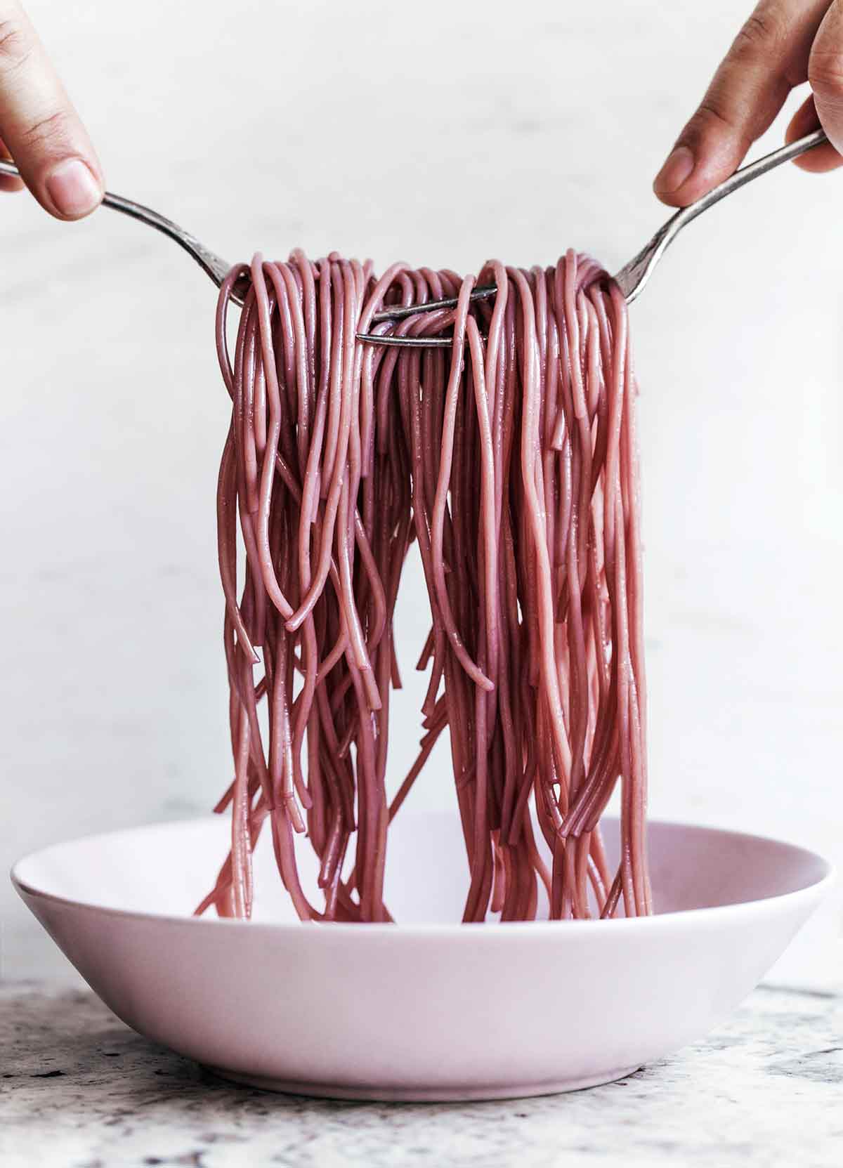 A person using two forks to lift strands of red wine spaghetti out of a white bowl.