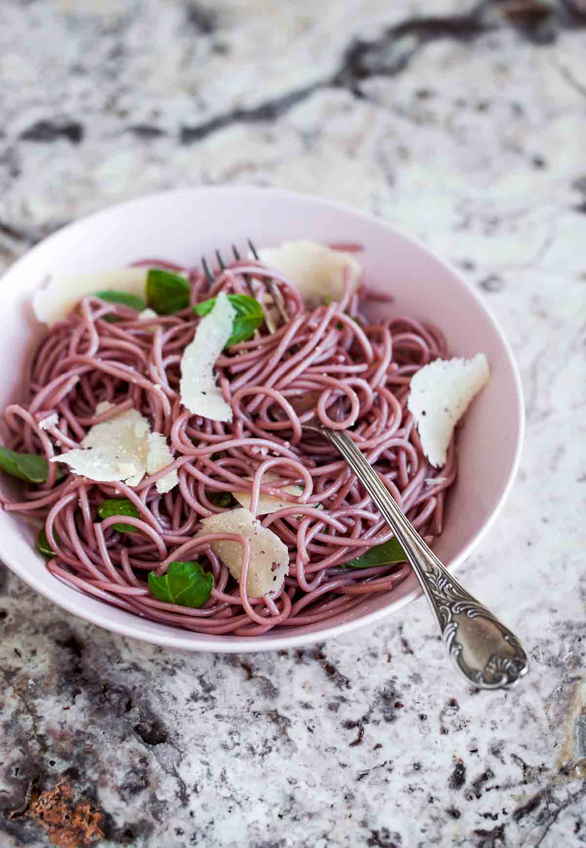A white bowl of red wine spaghetti with shaved Parmesan and basil leaves.