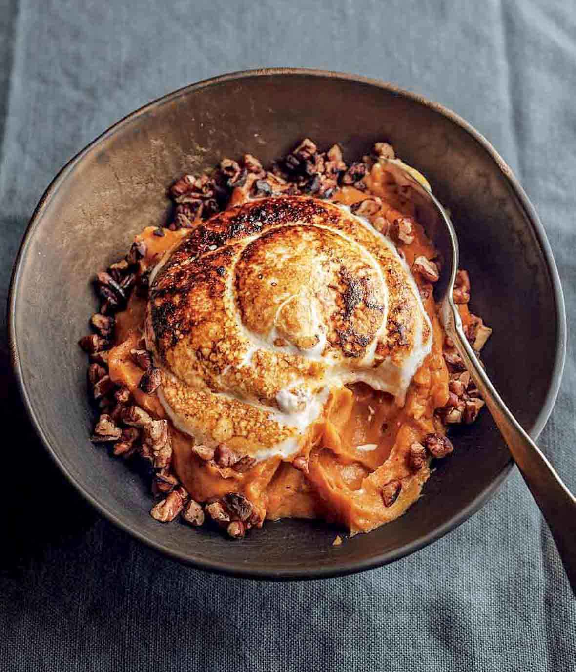 A bowl of sweet potato puree with marshmallow, which is toasted with a torch, toasted buttered pecans around the edge