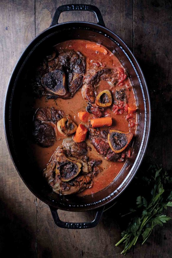 Five veal osso bucco, with carrots, and tomato sauce in. a black cast-iron pot.