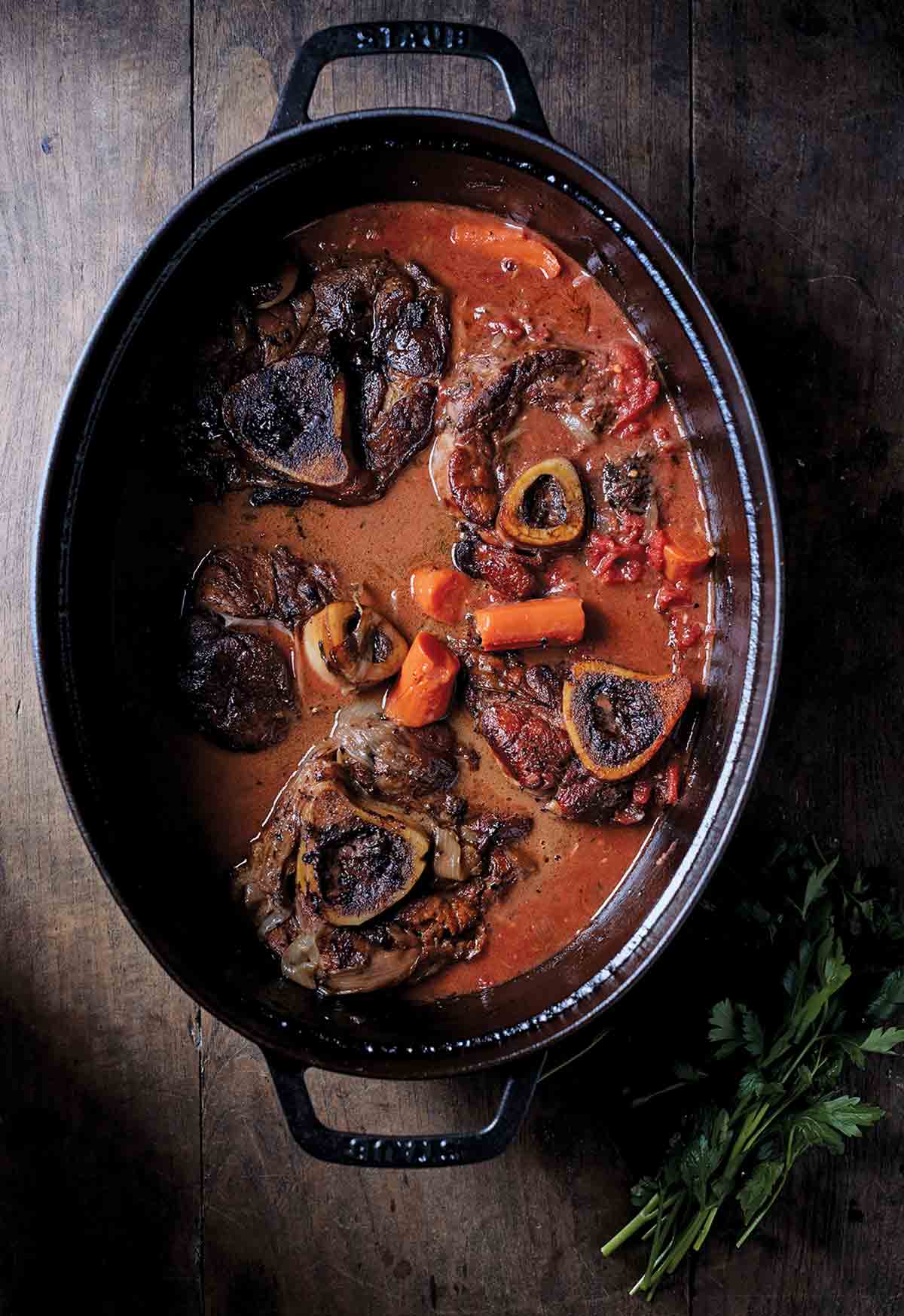 Five veal osso bucco, with carrots, and tomato sauce in. a black cast-iron pot.