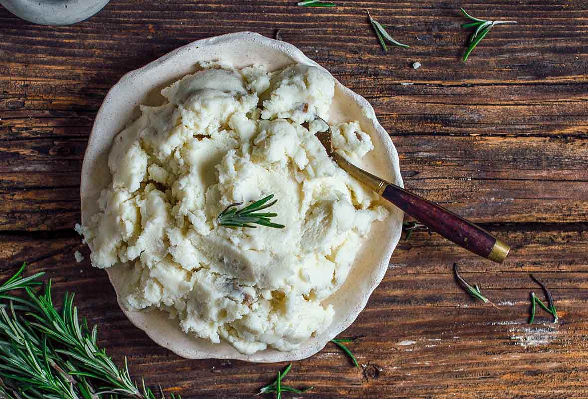 A white bowl of creamy vegan mashed potatoes with a sprig of rosemary and a spoon sticking in