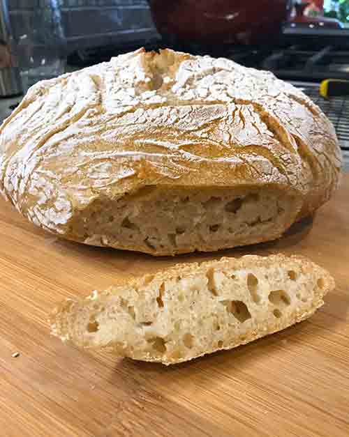 A loaf of Jim Lahey's no-knead bread on a cutting board with a slice removed; big air holes and a dusting of flour