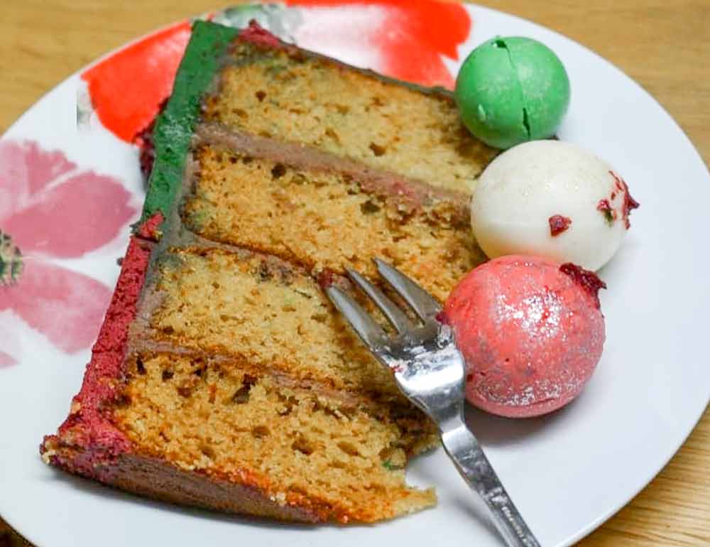 A slice of a three-layer vanilla paleo cake covered in red and green frosting and topped with candy globes