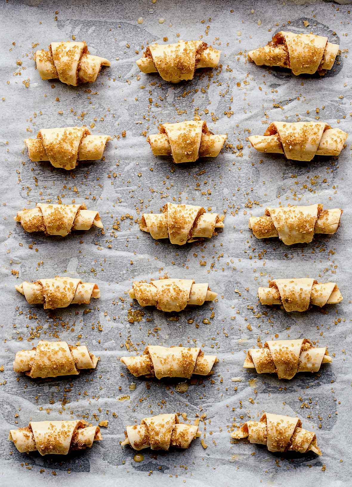 A baking sheet lined with parchment with 18 walnut rugelach cookies covered with brown sugar ready for the oven