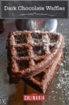 Two dark chocolate waffles on a mirrored surface sprinkled with powered sugar
