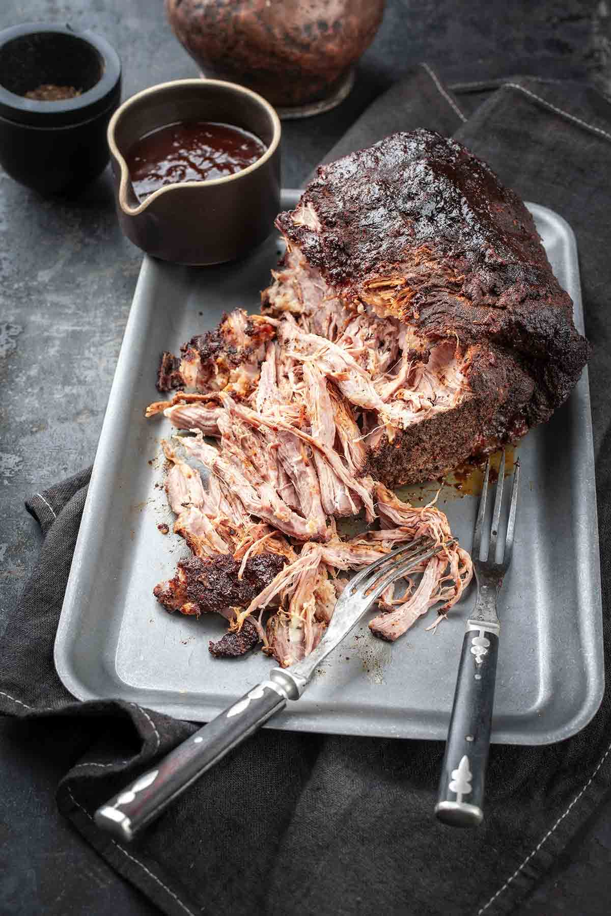 A partially shredded roast pork butt in a roasting pan with a cup of barbecue sauce and two forks.