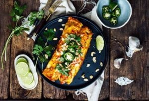 A casserole dish of sweet potato enchiladas with a place with two enchiladas, lime, jalapenos, and sauce