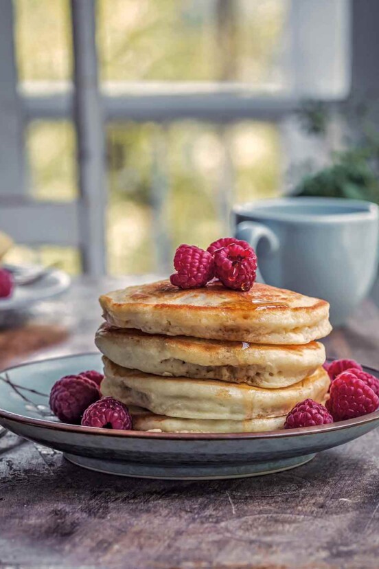 A stack of four vegan pancakes on a plate, dripping with maple syrup and topped with raspberries