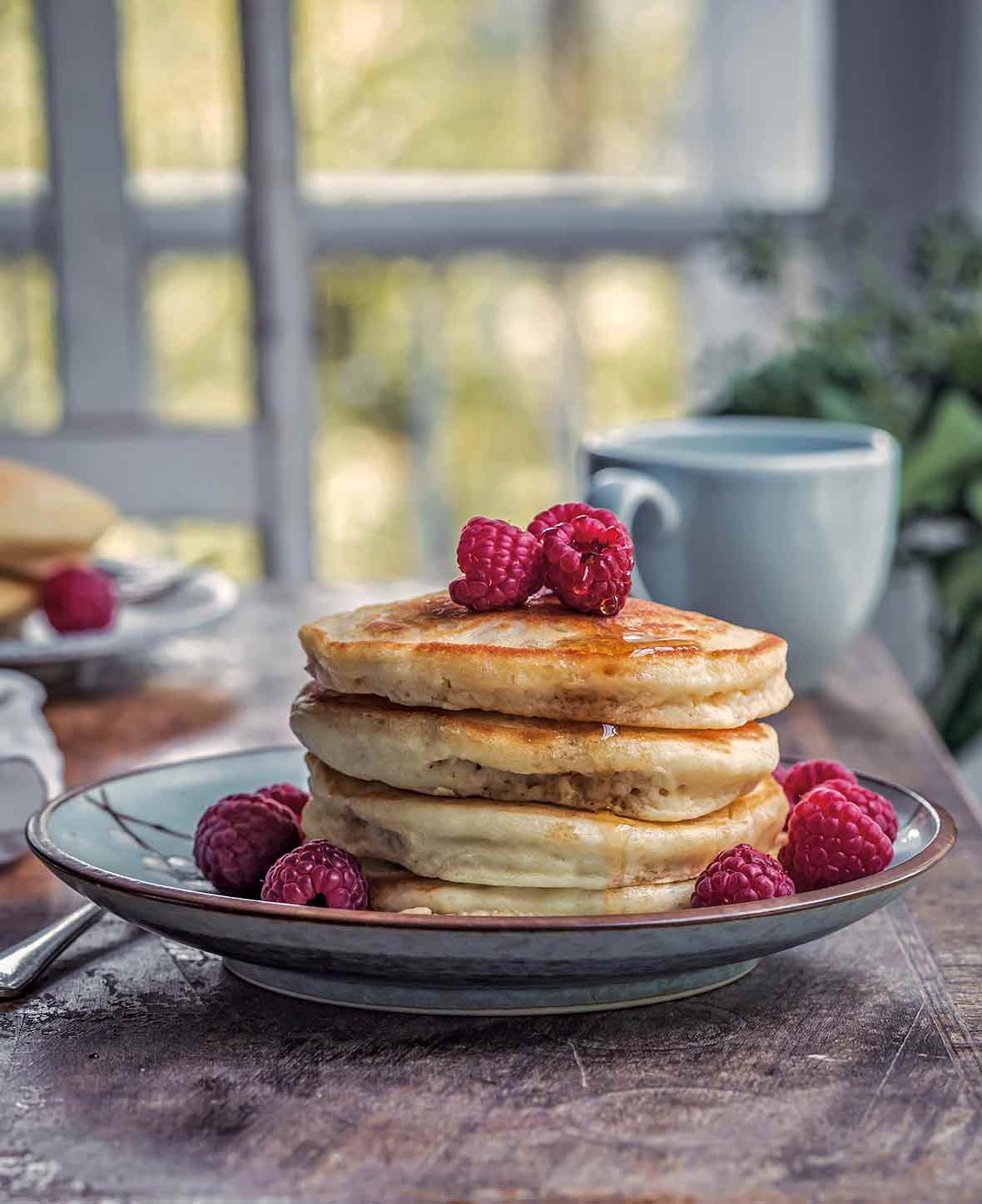A stack of four vegan pancakes on a plate, dripping with maple syrup and topped with raspberries