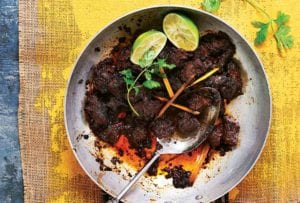 A stainless steel skillet filled with beef rendang and topped with two lime halves, lemongrass, and cilantro