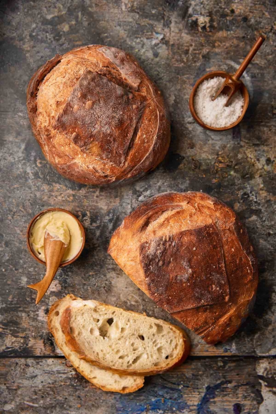 Two loaves of no-knead artisan bread, one with two slices cut from it and a dish of butter nearby.