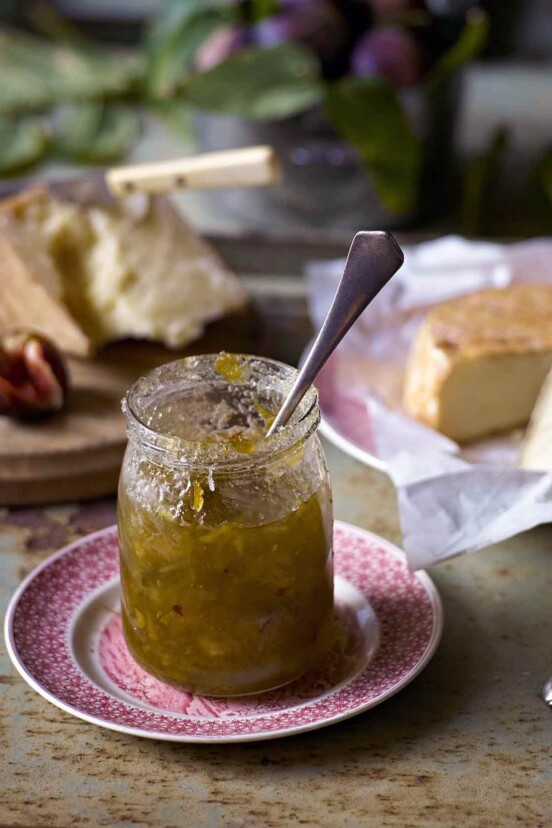 A jar of celery marmalade with a spoon in it and several types of cheese in the background