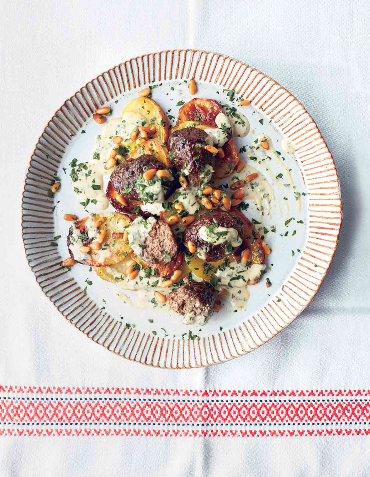 A white plate with striped edges filled with potatoes and lamb meatballs with tahini