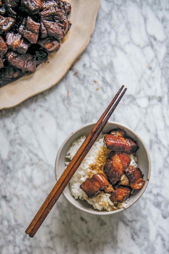 A bowl of rice topped with red cooked pork and a pair of chopsticks resting on the bowl. A plate of red cooked pork on the side