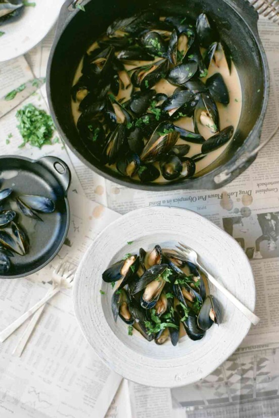 A pail and a white bowl filled with red curry mussels, as well as a bowl of discarded mussel shells