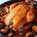 A best brined roast chicken in a cast iron Dutch oven surrounded by potatoes, mushrooms, carrots, and a halved head of garlic.