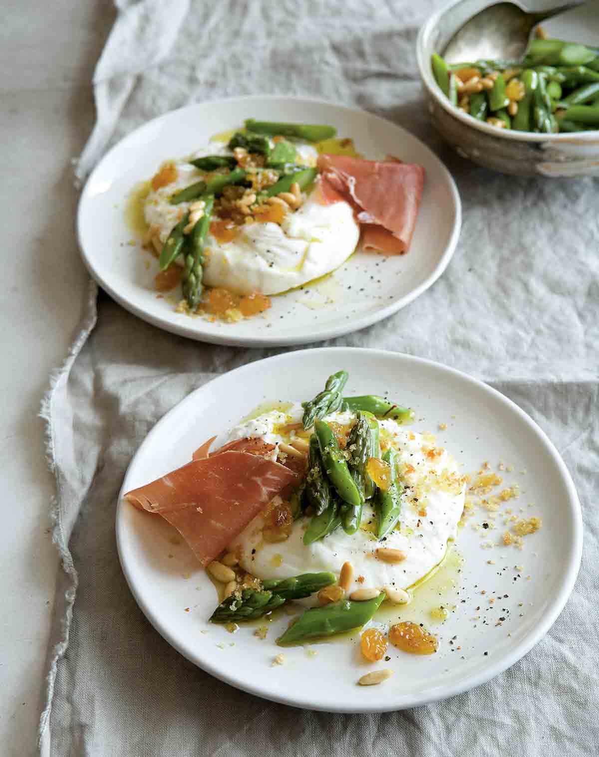 Two plates with burrata topped with asparagus, pine nuts, raisins, and prosciutto.