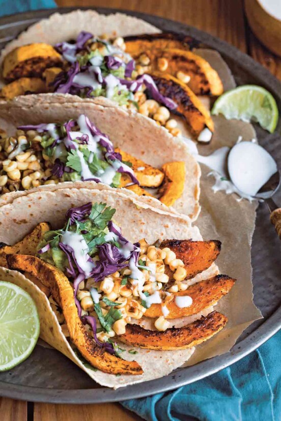 A platter of butternut squash tacos made with tortillas filled with strips of roasted squash, corn, cabbage, guacamole, and sour cream.