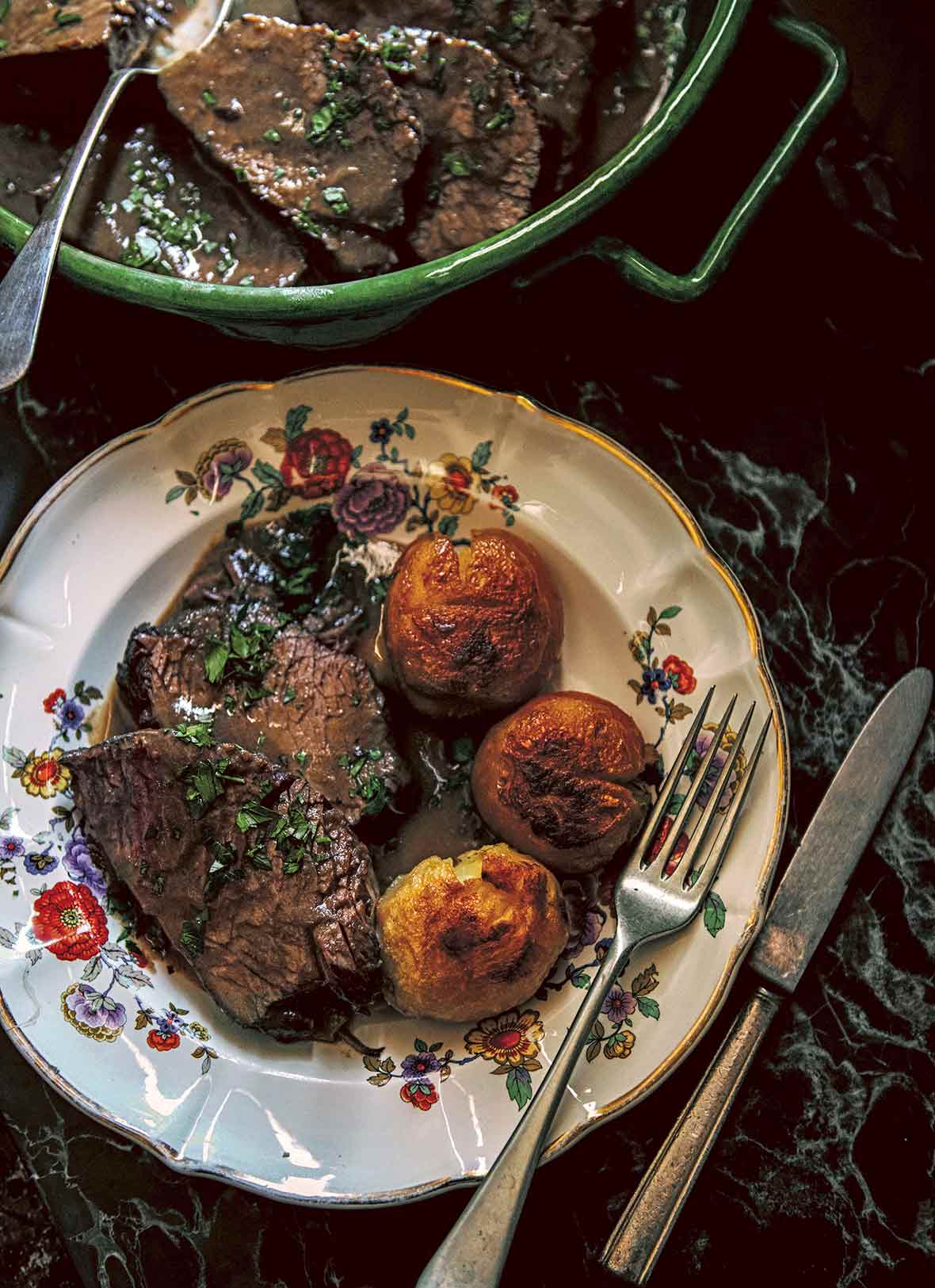 A china plate with three slices of beef braised in red wine and sprinkled with parsley, and three roasted potatoes