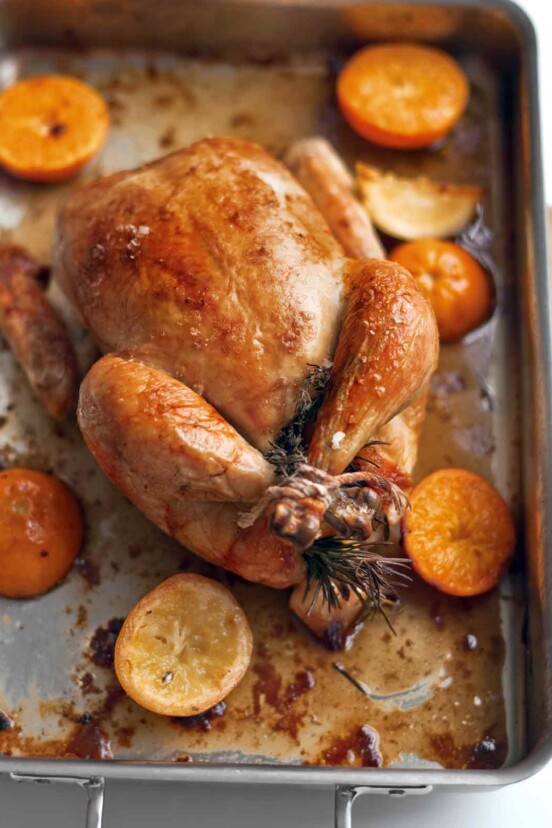 A whole roast chicken in a roasting pan with halved citrus fruits around it