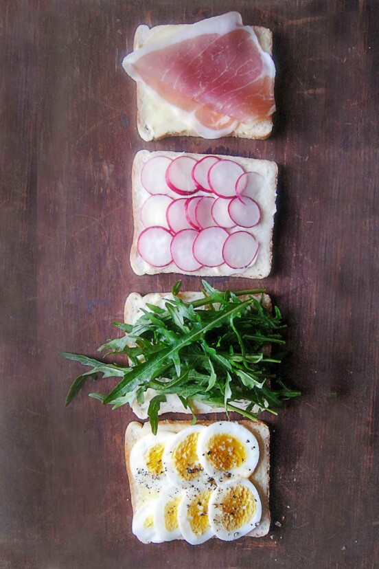 Tea sandwiches being assembled with four slices of bread topped with sliced egg, arugula, radish, and prosciutto