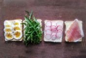 Tea sandwiches being assembled with four slices of bread topped with sliced egg, arugula, radish, and prosciutto