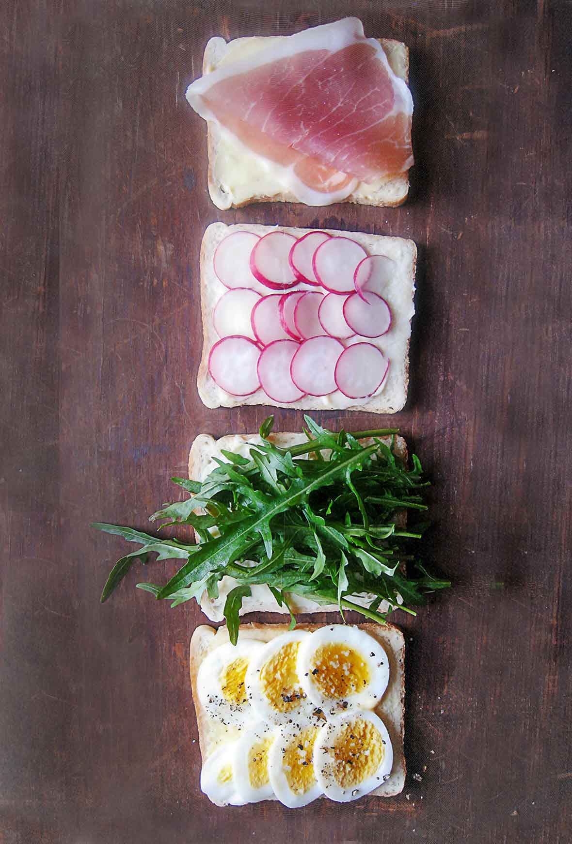 Tea sandwiches being assembled with four slices of bread topped with sliced egg, arugula, radish, and prosciutto