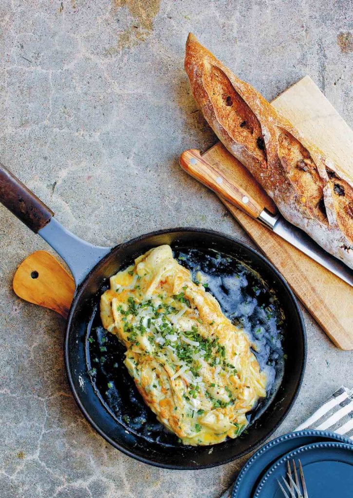 A black skillet containing a cheese omelet, topped with chives and Gruyere